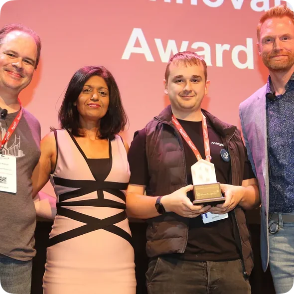 Four people stand on stage smiling, one holding an award trophy with a red backdrop reading Award.